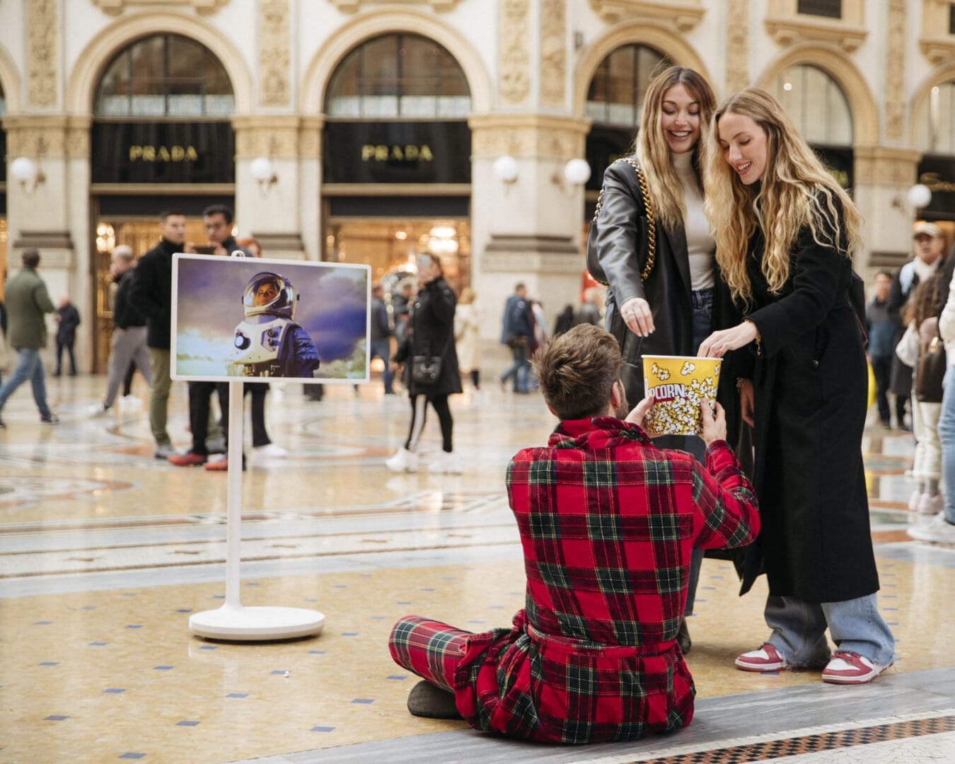 LG-adv-video-backstage-12 A person in plaid pajamas sits on the floor showing off his artwork, while holding out a yellow object to two smiling women, in a scene reminiscent of a fast-paced fashion movie.