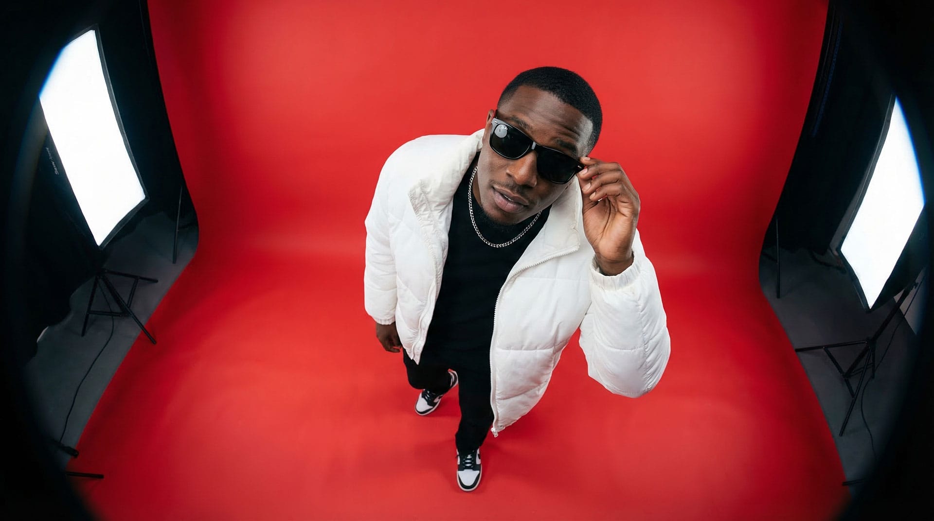 examples-images-ai-7 A person in a white jacket and sunglasses stands against a red backdrop, ready for a Reel photo shoot, with studio lights on either side.